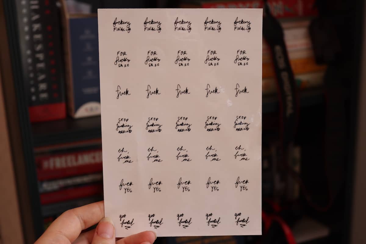 An image of a person holding a sticker sheet with small, round transparent stickers that say curse words, like "fuck" in front of a bookshelf.